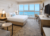 Hotel room with a large bed, a wall-mounted TV, armchair, and a view of the ocean through a wide window.