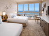 Hotel room with two beds, a dresser, and a large window overlooking a marina with boats and the ocean in the background.