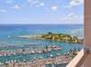 View from a balcony overlooking a marina with docked boats, a small peninsula with trees and grass, and the ocean extending to the horizon under a clear sky.