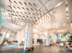 Modern hotel lobby with white pillars, a decorative ceiling feature, and seating areas. Large windows allow natural light to illuminate the spacious room.