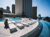 A rooftop infinity pool with surrounding empty lounge chairs and a city skyline in the background on a clear day.