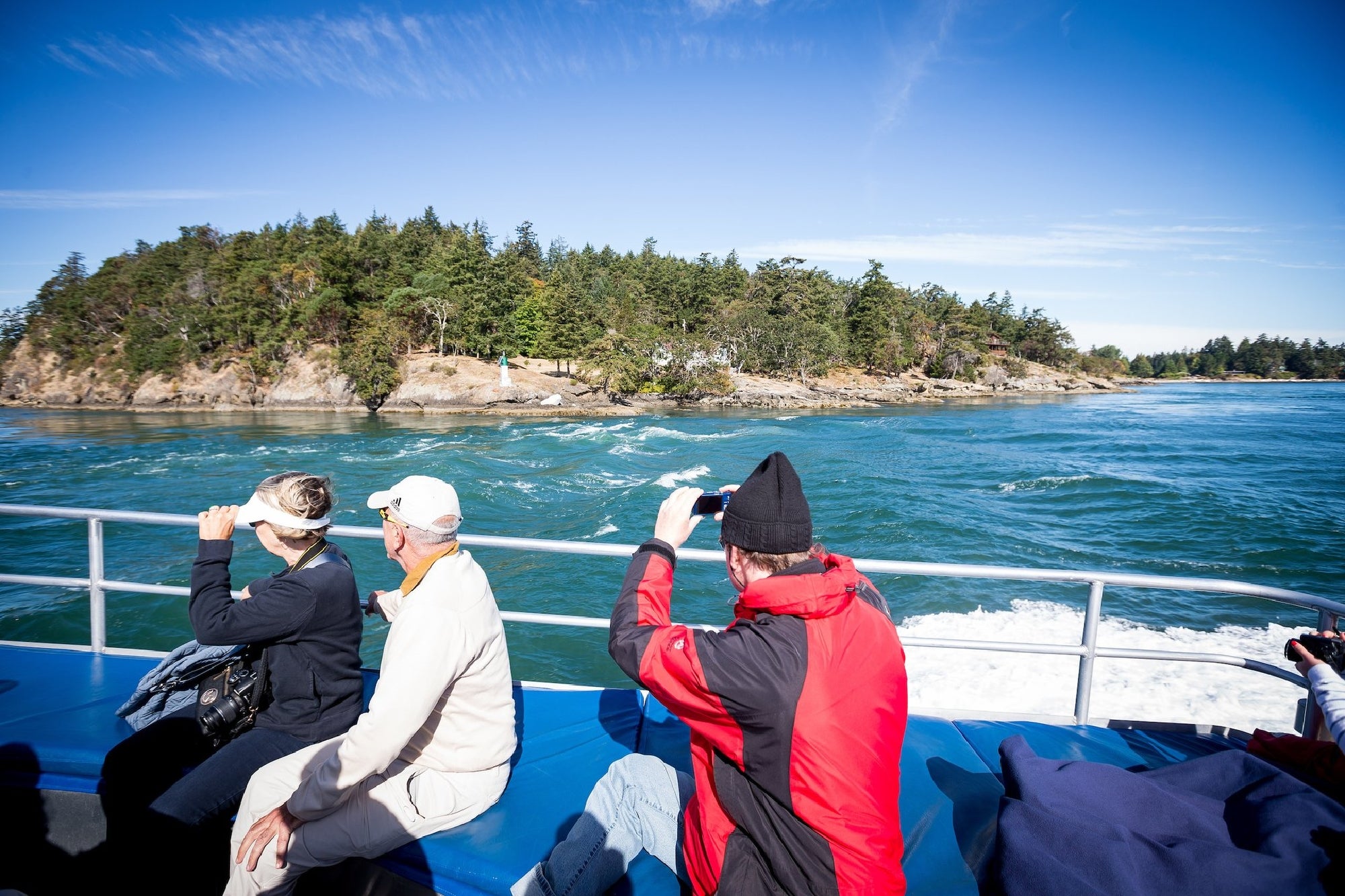 People on a boat take photos of an island with trees under a clear blue sky.