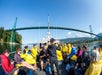 A group of people wearing yellow raincoats sit on a boat under a large suspension bridge with green towers, surrounded by green hills and water on a sunny day.