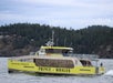 A yellow and gray catamaran named "Prince of Whales" moves through water with a wooded shoreline in the background.