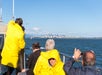 People in yellow jackets on a boat look towards a distant city skyline under a clear blue sky.