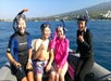 Four people in snorkeling gear sitting on a boat, smiling, with a coastline and a mountain in the background.