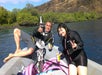 Two people sitting on a boat, smiling, holding drinks, with a scenic backdrop of water, trees, and rocky hills. Pink flotation noodles are visible in the foreground.