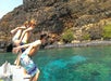 Three boys in swim trunks are about to jump off a boat into clear blue water, with a rocky, tree-dotted shoreline in the background.