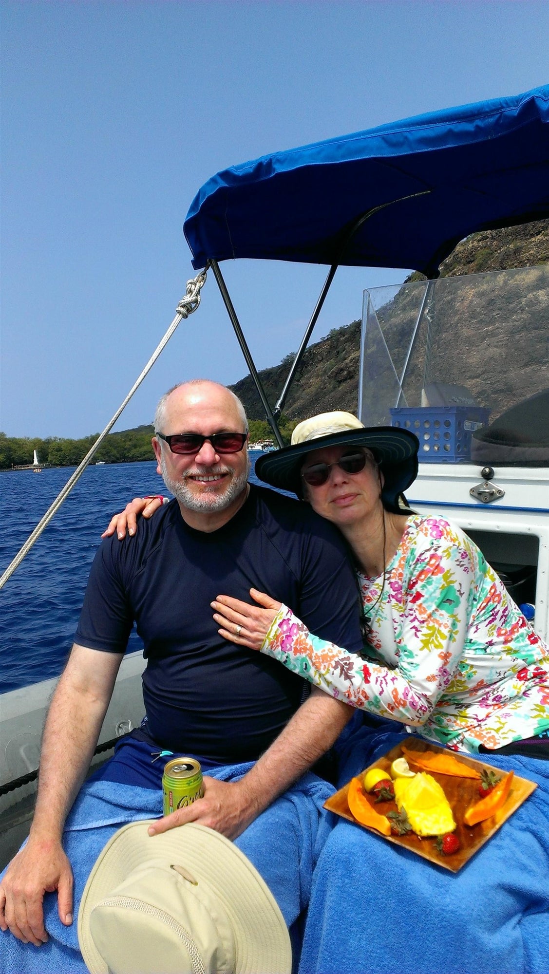 Couple seated on a boat under a blue canopy, with a man holding a can and a woman wearing a sunhat. She's resting her hand on his shoulder. A plate of snacks is beside them.