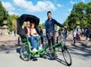 Two people sit in the back of a green pedicab while a driver stands beside it, outdoors in a park with trees and people in the background.