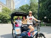 A woman pedals a blue rickshaw with two smiling passengers seated in the back on a sunny day in an urban park.