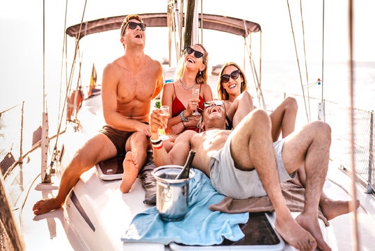 Four young adults sit and lounge on a sailboat deck, laughing and holding drinks, with a bottle of champagne in an ice bucket nearby and the ocean in the background.