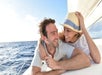 A smiling man and woman relax together on a sailboat at sea, with the woman wearing a hat and leaning toward the man. The ocean and blue sky are visible in the background.