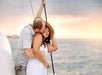 A man embraces and kisses a smiling woman wearing a hat. They are standing on a boat with the ocean and a sunset in the background.