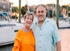 Two adults stand close together and smile at the camera, with boats and water in the background at what appears to be a marina.
