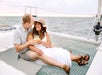 A couple sits close together on a catamaran net, smiling and holding drinks, with the ocean and sky in the background.