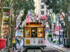 A cable car travels down a bustling urban street lined with trees and shops, as people walk on the sidewalks.