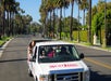 Tour bus driving through a palm-lined street, carrying passengers seated in the open section at the back.