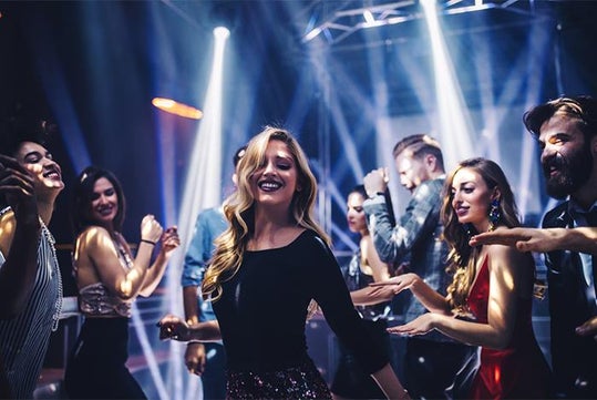 A group of young adults dancing and smiling under colorful lights in a nightclub setting.
