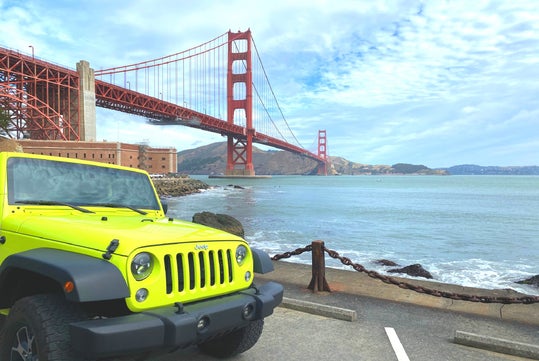 A bright yellow Jeep is parked by the waterfront with the Golden Gate Bridge and hills visible in the background under a cloudy sky.