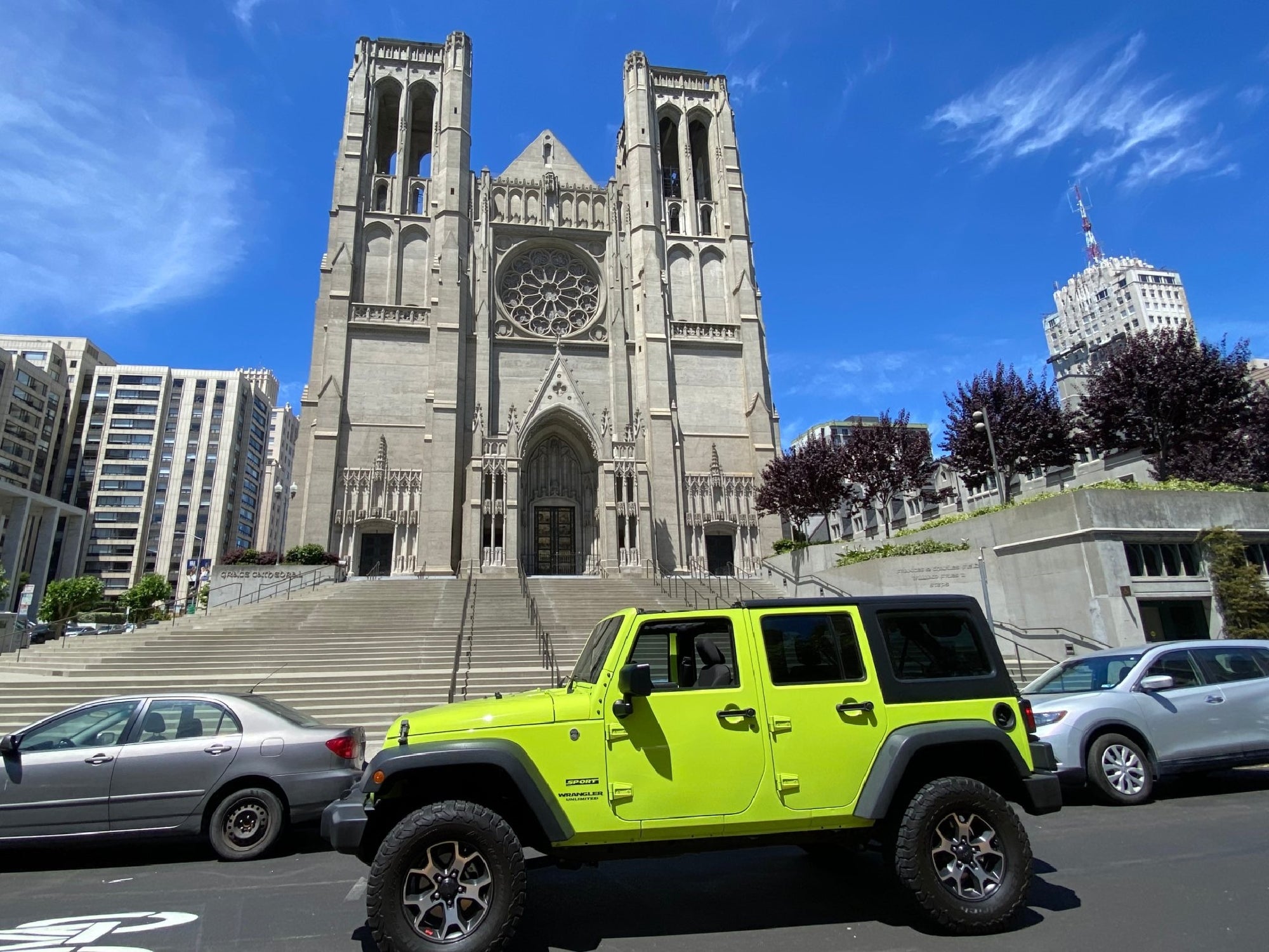 A bright green Jeep is parked on a street in front of a large stone cathedral with tall towers and a rose window, surrounded by other city buildings.