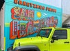 A bright yellow Jeep parked in front of a colorful mural that reads “Greetings from San Francisco” with city landmarks illustrated inside the letters.