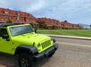 A bright green Jeep is parked on a street in front of a row of large, red-roofed brick buildings under a partly cloudy sky.