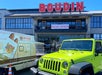 A Boudin bakery with a "Now Open" sign above the entrance, a delivery van, and a bright green Jeep parked in front.