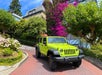 A bright green Jeep Wrangler parked on a red brick road, surrounded by trees, flowers, and houses on a sunny day.