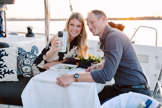 A woman and man sit at a table on a boat, smiling and taking a selfie together with a smartphone during sunset.