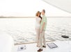 A woman and man stand together on the deck of a boat, smiling and looking at each other, with calm water and distant shoreline in the background.