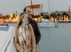 A couple stands together on a boat, smiling, with water, yachts, and illuminated buildings in the background at dusk.