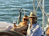 Two people sit on a sailboat, smiling at the camera and holding glasses of red wine, with the ocean visible in the background.