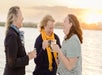 Three women standing by the water at sunset, holding drinks and smiling while talking together.