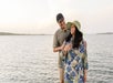 A man and woman stand together by the water, smiling. The man wears a cap and a short-sleeve shirt; the woman wears a sun hat and a floral dress. The background shows a calm lake.