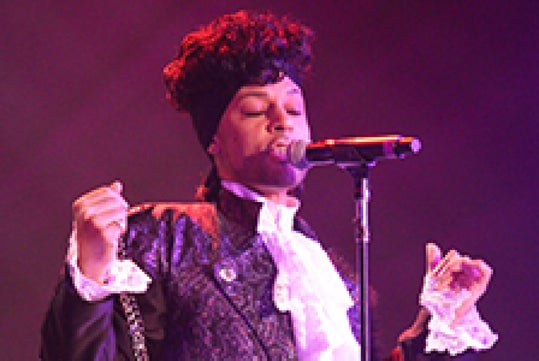 A Prince impersonator singing into a microphone wearing a purple jacket with a dark purple light shining on the stage.