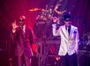 Two men performing on stage wearing suits and sunglasses with a background lit up with pink and purple lights.