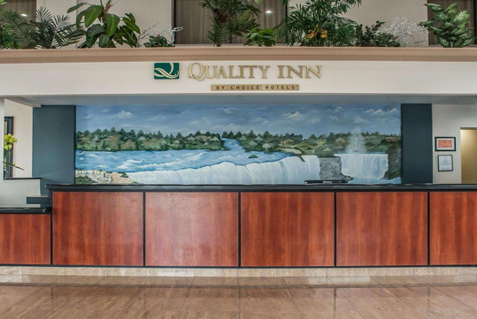 Hotel front desk area with a large mural of a waterfall behind the counter, under a sign that reads "Quality Inn by Choice Hotels.