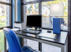 A desk by a window with a computer, keyboard, mouse, printer, and a blue speaker. An empty blue chair is at the desk.