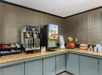 A breakfast buffet with coffee machines, a juice dispenser, fruit basket, oatmeal packets, and condiments on a granite countertop against a textured wall.