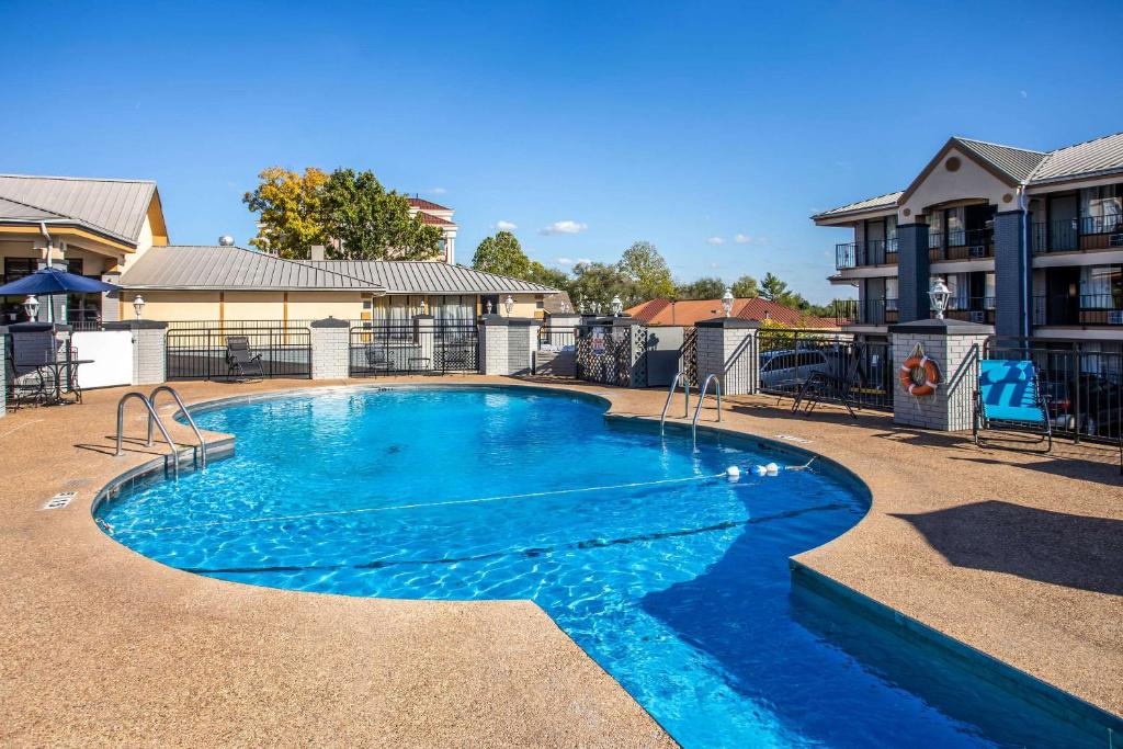 Outdoor swimming pool with clear blue water, surrounded by a brown deck. Three-story buildings with balconies and a blue sky are in the background.