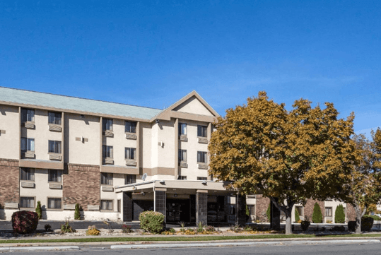 Four-story hotel building with beige and brown exterior. Large tree with autumn leaves on the right. Clear blue sky overhead.