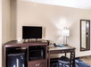 Hotel room corner with a TV on a wooden cabinet, a mini-fridge, a microwave, a desk with a lamp, a chair, and a mirror on the wall.