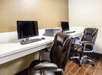 Two office chairs at a desk with two computers and a printer in a room with beige walls and tiled floor.