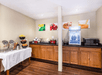 A small kitchen area with wooden cabinets, a counter, microwave, mini fridge, and breakfast items. Art on walls.