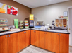 Hotel breakfast area with cereal dispensers, bread, pastries, coffee urns, juice machine, and fruit display under colorful fruit-themed wall art.