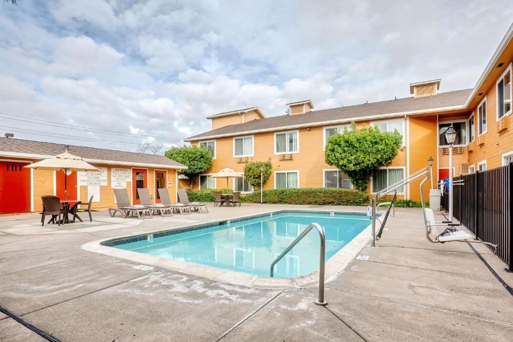 Outdoor swimming pool area with loungers, umbrellas, and a fence, adjacent to a two-story orange building under a cloudy sky.