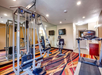 A small gym room with a treadmill, weight machine, exercise bike, mirrors, and two wall-mounted TVs. The floor has a colorful patterned carpet.
