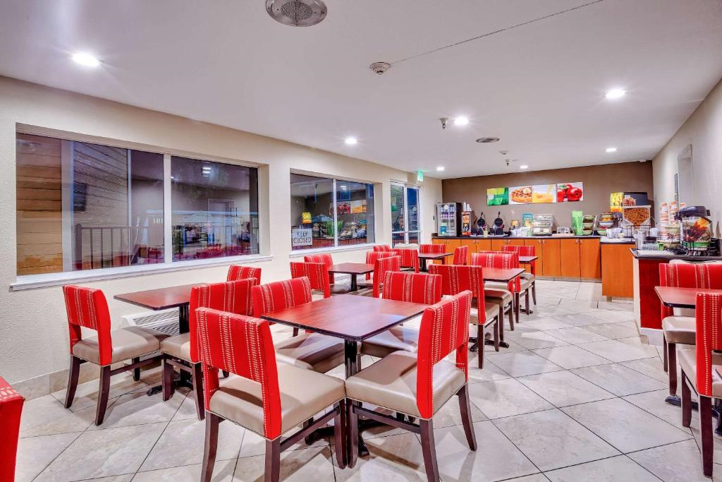 A brightly lit hotel dining area with red upholstered chairs and wooden tables. The room has tiled floors, large windows, and a breakfast buffet setup along the back wall.