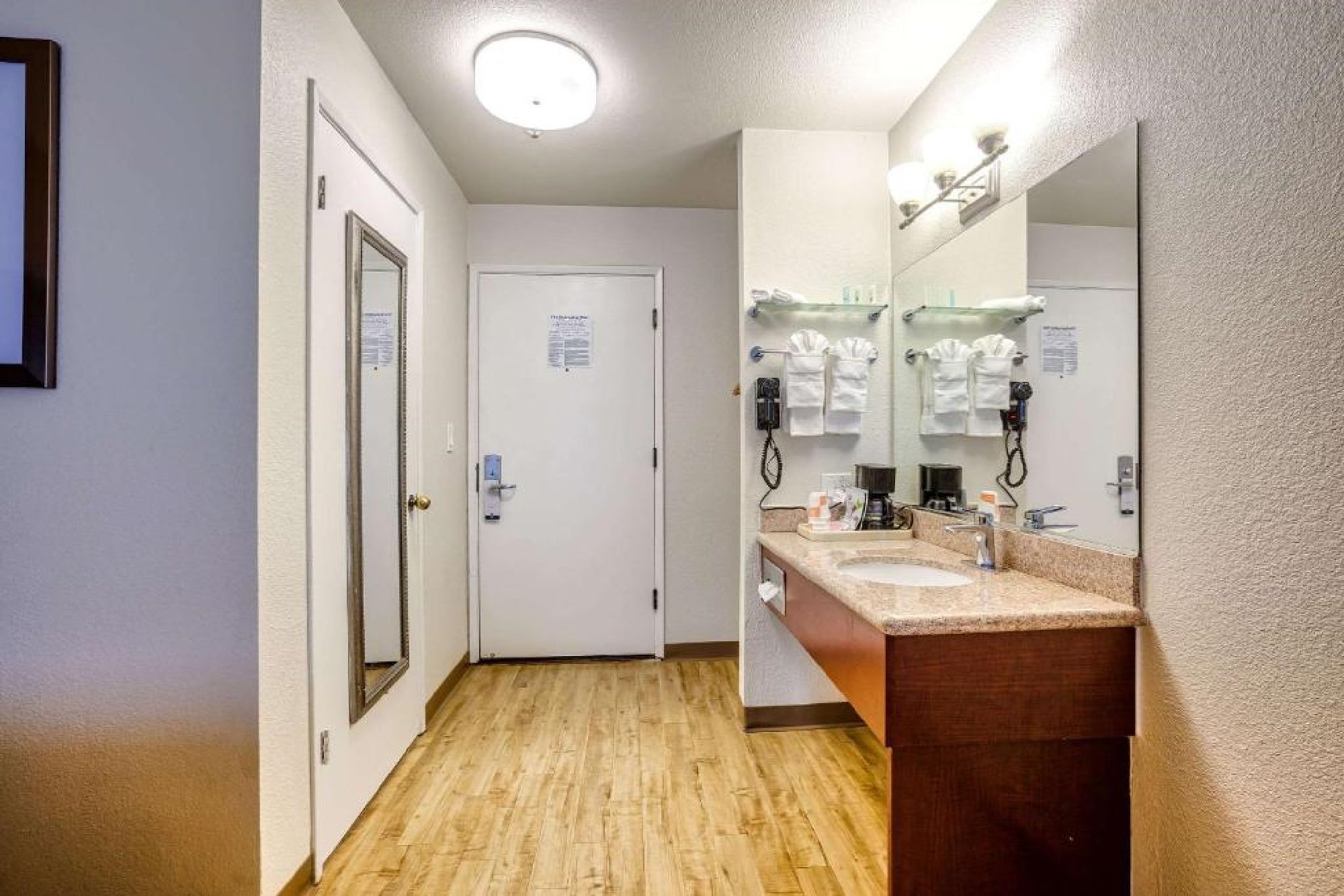 Hotel bathroom with a wooden floor, large mirror, sink, and toiletries. Wall-mounted hairdryer and towels are neatly arranged. Door to the room is visible.